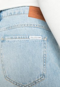 Ljusblå denimjeans med en texturerad yta. Har en bakficka med en vit etikett som läser "Marc O’Polo DENIM" och en brunt läderpatch på midjan.