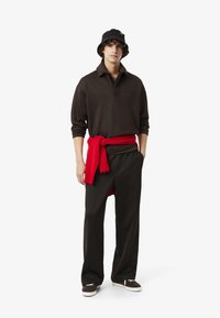 Ung man som står iförd svart bucket hat, mörk långärmad polo, svarta vida byxor, svarta skor och en röd tröja knuten runt midjan.