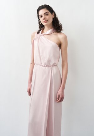 Młoda kobieta o ciemnych, kręconych włosach, ubrana w bezrękawową, jasno-różową sukienkę na jedno ramię, stojąca na tle jasnego, jednolitego tła.