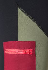 Detailní záběr na černou a olivovou tkaninu s červenou kapsou na zip, která má jasně červený plastový táhlo zipu.