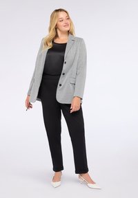 Donna bionda che indossa una maglietta e pantaloni neri, con un blazer grigio a scacchi e tacchi bianchi a punta, in piedi su uno sfondo semplice.