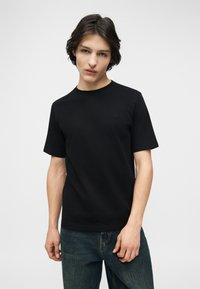 Νεαρός άνδρας με μεσαίου μήκους σκούρα μαλλιά, φορώντας απλό μαύρο κοντομάνικο πουκάμισο και μπλε τζιν, μπροστά σε λευκό φόντο.