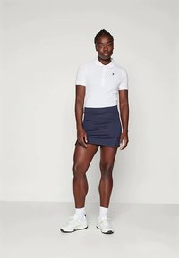 Sportos nő áll, fehér pólót, tengerészkék sport szoknyát oldalsó hasítékokkal, fehér zoknit és fehér tornacipőt visel, egy sima háttér előtt.