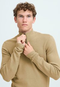 Maglione beige con zip al collo, realizzato in un morbido tessuto testurizzato. Presenta polsini e colletto a costine, offrendo una forma aderente lungo tutto il corpo.