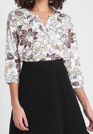 Vrouw draagt een witte bloemenblouse met driekwart mouwen, ingestopt in een gestructureerde zwarte A-lijn rok.
