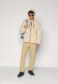 Kreemikas fliisjope kapuutsiga, ees lukuga ja kontrastse äärega, kantud beežide pükste ja mustade kingadega, kaasas sinine seljakott.