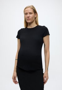 Ξανθιά γυναίκα με εφαρμοστή μαύρη κοντομάνικη μπλούζα και μαύρη φούστα, στέκεται με ουδέτερη έκφραση μπροστά σε απλό λευκό φόντο.