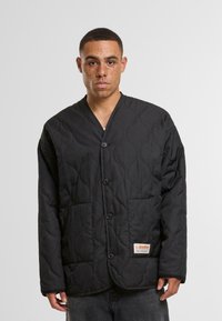 Black quilted jacket with a mandarin collar, featuring button front closure and two front pockets. Label on left pocket.