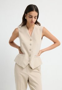 Gilet beige sans manches avec un décolleté en V profond, cinq boutons, deux poches latérales et un design structuré. Tissu lisse avec une coupe sur mesure.