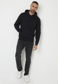 Un hoodie noir avec une poche kangourou et une capuche à cordon, associé à un jean gris foncé et des baskets blanches. Le hoodie présente un petit logo sur la poitrine.