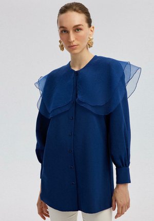 Chemise bleue avec un grand col transparent superposé, des manches longues et un devant boutonné. Fabriquée dans un tissu lisse et léger avec une coupe légèrement ample.