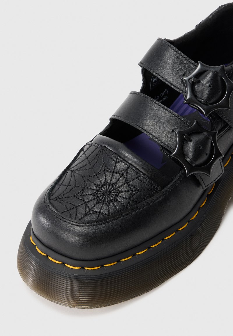 Chaussure plateforme en cuir noir avec broderie en toile d'araignée sur le bout, deux sangles avec boucles rondes à pointes, et coutures jaunes sur la semelle.