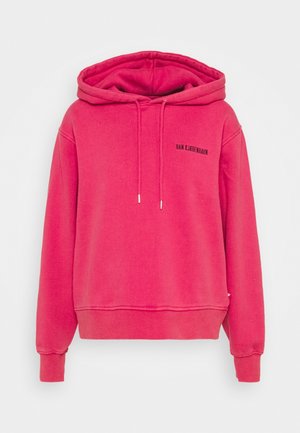 Henkilö, jolla on päällään kirkkaanpinkki huppari, musta nahkainen nappikiinnitteinen minihame ja mustat nilkkurit valkoista taustaa vasten.