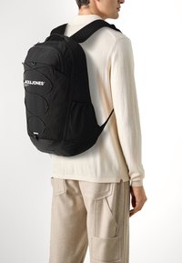 Black backpack featuring smooth material, adjustable straps, logo detail, and elastic cord on front. Positioned against a light background.