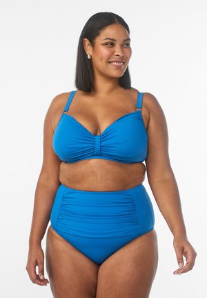 Smiling woman with medium skin tone wearing a blue two-piece swimsuit with ruched details, standing against a plain white background.
