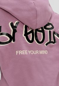 Un sweat à capuche violet en tissu doux, avec un grand texte noir de style graffiti dans le dos portant l'inscription "LIBÈRE TON ESPRIT".