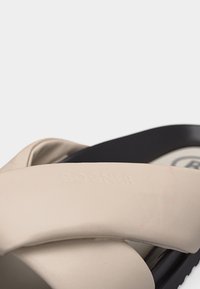 Gros plan sur les brides de sandales beige rembourrées croisées sur une semelle noire avec le nom de la marque embossé sur la bride.