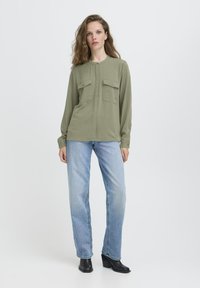 Femme aux cheveux ondulés portant une chemise à manches longues vert olive avec des poches à l'avant, un jean bleu clair et des bottines à talons noires, debout.