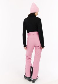 Vrouw in roze skibroek, zwart geribbeld shirt met lange mouwen, roze gebreide beanie en zwarte skischoenen, van achteren gezien tegen een witte achtergrond.