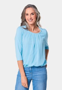 Hellblaue Bluse mit dreiviertel Ärmeln, gefurchter Vorderseite und lockerer Passform. Kombiniert mit Jeans, vor einem schlichten Hintergrund gezeigt.