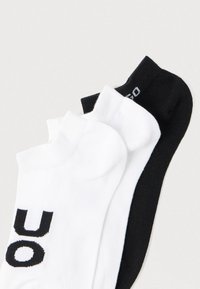 Τρία ζευγάρια κάλτσες αστραγάλου: δύο λευκές με μαύρα γράμματα και μία ολόμαυρη. Μείγμα βαμβακιού, με ριμπ μανσέτες και λεία υφή.