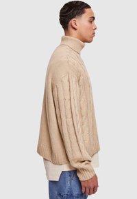 Pulover bej tricotat cu guler înalt, având modele din tricot cu cabluri și manșete drepte. Purtați peste o cămașă de culoare deschisă, prezentat din lateral.