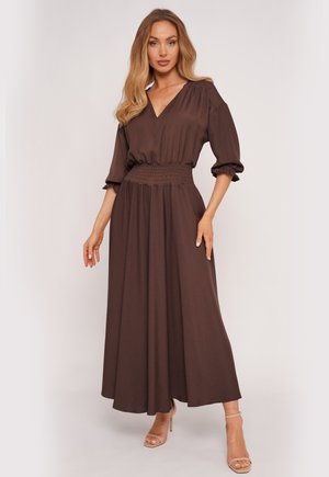 Kobieta o długich blond włosach, ubrana w długą brązową suknię z dekoltem w szpic, rękawami 3/4 i marszczonym pasem, stojąca w beżowych sandałach na wysokim obcasie.