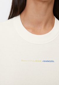 Nærbillede af en person iført en hvid ribbet t-shirt med rund hals og gul og blå "Marc O'Polo DENIM x KANGOL" tekst på brystet.