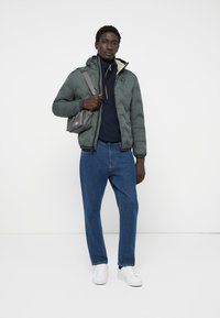 Padded green jacket with a zip, navy sweater, blue jeans, and white sneakers. Model holds a small gray bag with a strap.