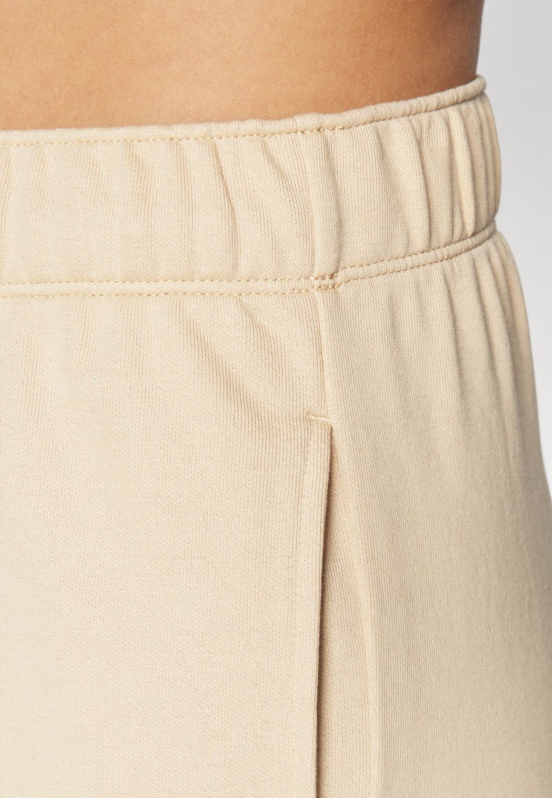 Tissu extensible beige avec une taille élastique, présentant une couture latérale et une texture lisse. Un angle rapproché met en évidence les détails de couture.