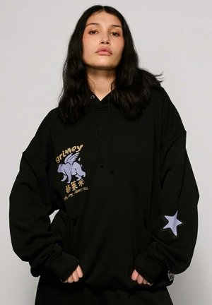 Jonge persoon met lang donker haar, gekleed in een zwarte hoodie met een afbeelding van een gevleugelde griffioen, gouden tekst en een grijze ster op de mouw.