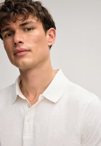 Witte polo met een textuurpatroon, korte mouwen en knoopsluiting. De kraag is klassiek en iets losgeknoopt.