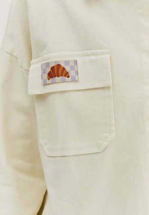 Beige košile s kapsou a kostkovaným látkovým záplatou, na které je stylizovaný design oranžového croissantu. Viditelná struktura látky.