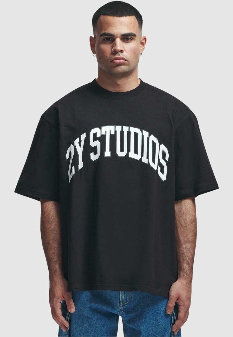 Black cotton t-shirt with oversized fit, featuring white "ZY STUDIOS" text in an arched design across the chest. Short sleeves, crew neck.