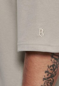 Vaaleanharmaa lyhythihainen t-paita, jossa on pieni brodeerattu "B" -logo hihassa, valmistettu pehmeästä puuvillakankaasta, jolla on sileä rakenne.