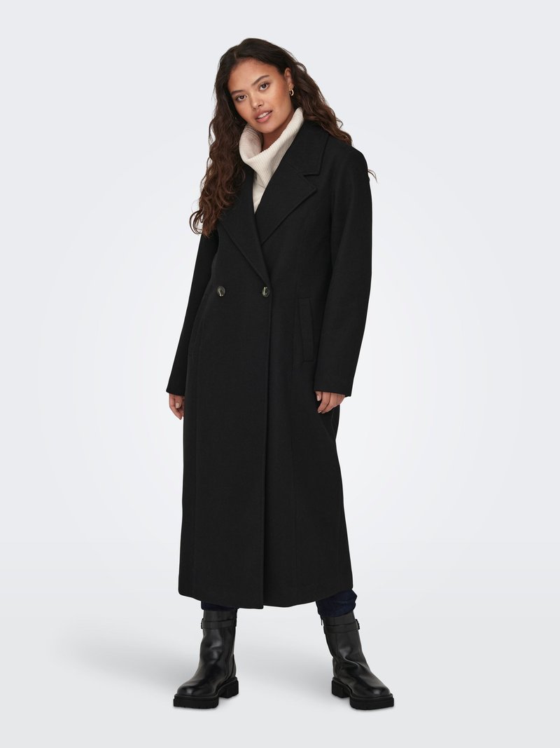 Vrouw draagt lange zwarte dubbelgeknoopte jas, crème coltrui, donkere broek en zwarte enkellaarsjes, staand op witte achtergrond.