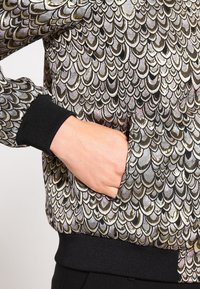 Metaliczna kurtka bomber z wzorem łusek ryb w kolorach złotym, srebrnym i czarnym, z prążkowanymi mankietami i kieszeniami bocznymi. Teksturowane wykończenie.