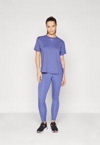 Lilla treningstrøye med korte ermer og sidespalter, kombinert med matchende lilla leggings. Svarte joggesko fullfører antrekket.