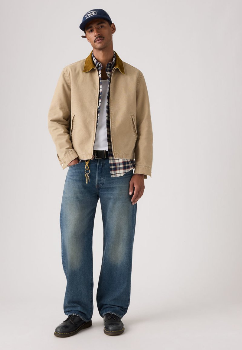 Chaqueta beige con cuello de pana marrón sobre una camisa a cuadros, combinada con vaqueros sueltos azules y zapatos negros, posando frente a un fondo neutro.