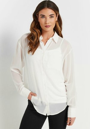 Camicia bianca a maniche lunghe, con bottoni, realizzata in un tessuto leggero, dotata di una singola tasca sul petto e orlo curvo. Abbinata a pantaloni scuri.