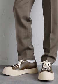 Zapatillas de piel marrón con suela de goma blanca y cordones. Detalles notables incluyen ojales y un diseño de puntera redondeada. Llevadas con pantalones de color tostado.