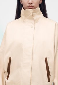 Κοντινό πλάνο ενός ατόμου που φορά ένα ανοιχτό μπεζ μπουφάν με ψηλό γιακά και κάθετες τσέπες με καφέ δερμάτινες επενδύσεις.