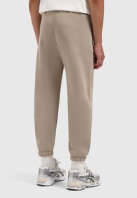 Beige joggingbroek met elastische manchetten en zijzakken, gemaakt van zachte stof; gedragen met grijze en witte sportschoenen.