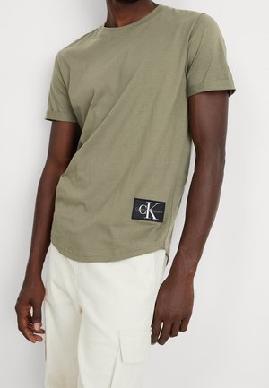 Mężczyzna w oliwkowej koszulce z krótkim rękawem z naszywką z logo Calvin Klein Jeans oraz białych spodniach z boczną kieszenią na białym tle.