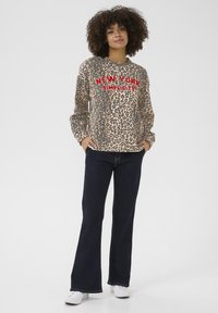 Mikina s leopardím vzorom v béžovej a hnedej farbe s červeným nápisom „NEW YORK SIMPLICITY“; skombinovaná s rozšírenými tmavými džínsami a bielymi teniskami.
