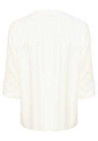 Blouse blanche à manches longues avec poignets retroussés, coupe ample et détail de tissu froncé dans le haut du dos.