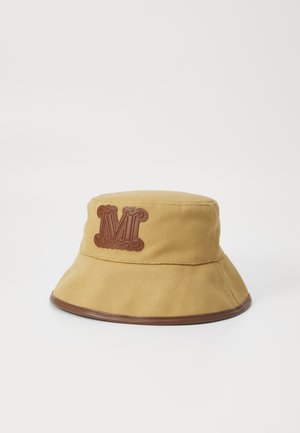 Chapeau seau en tissu texturé jaune moutarde, avec un logo en cuir brun en forme de "M" et un bord brun à la base.