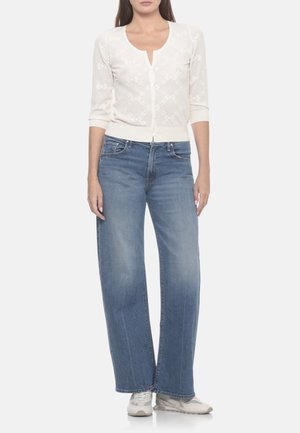 Donna che indossa un cardigan bianco a motivi con maniche a tre quarti, jeans blu larghi e sneakers bianche, in piedi su sfondo bianco.