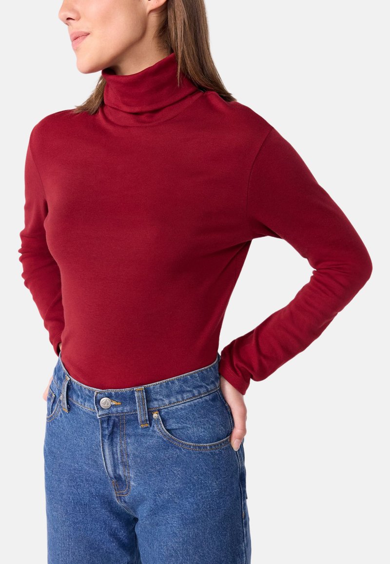 Maglione rosso a maniche lunghe con collo alto realizzato in tessuto morbido, abbinato a jeans in denim blu a vita alta con chiusura a bottone e tasche anteriori.