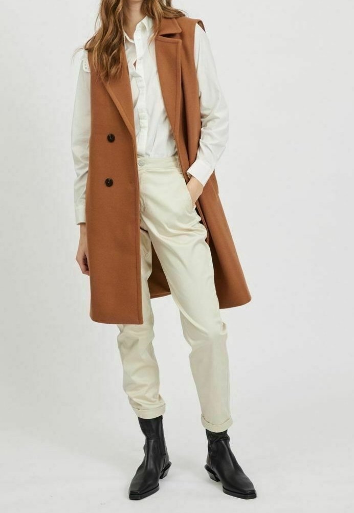 Femme portant une chemise blanche boutonnée, un pantalon beige taille haute, un manteau sans manches marron et des bottines noires, debout avec une main dans la poche.